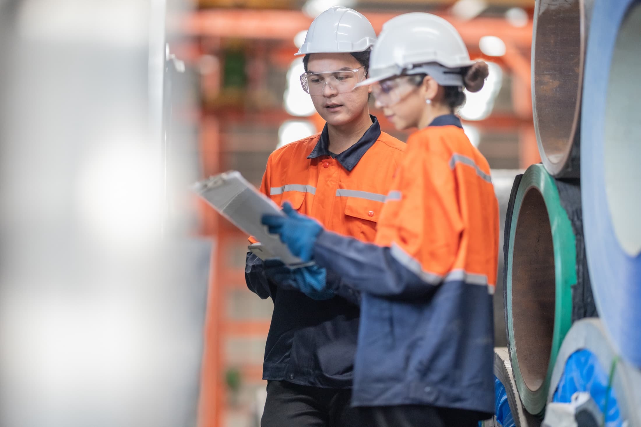 Two inspectors in high-visibility safety gear examining a clipboard at a metal-product manufacturing site.