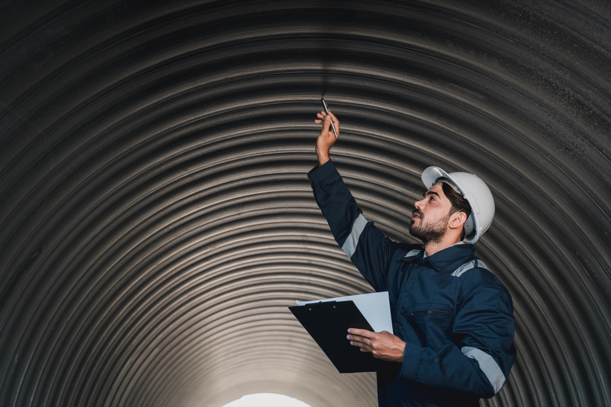 Inspector in navy coveralls and white hard hat examining the upper surface of a curved corrugated structure with a clipboard.