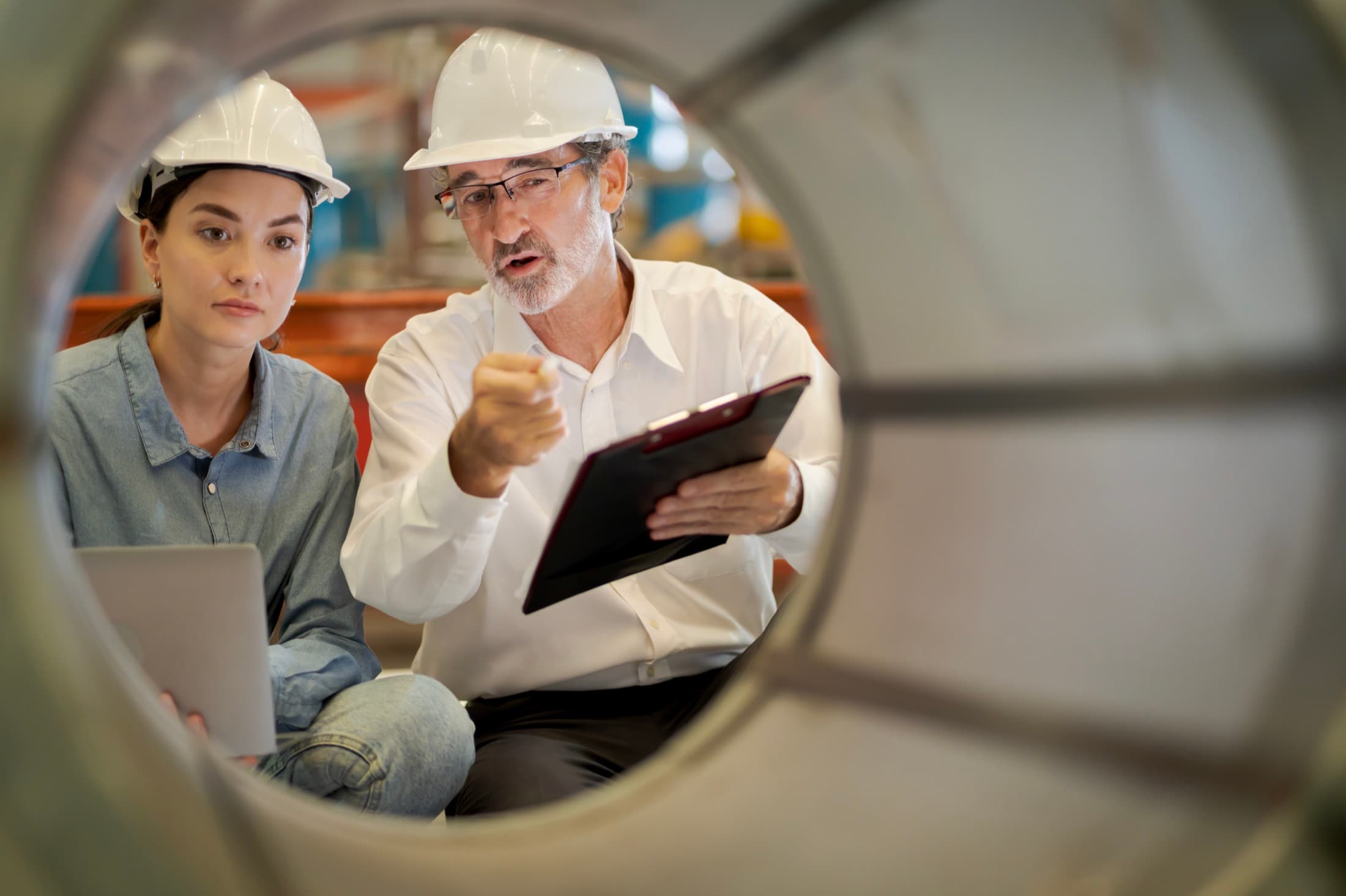 Two inspectors in hard hats reviewing a checklist on a tablet, framed by an industrial pipe section — collaboration inside the managed network.
